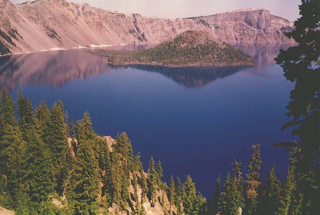 This photograph, also taken from the west rim looks out over the lake and shows Wizard Island. Wizard Island is actually the 'crater' of Crater Lake. You can see the crater in the top of the cinder cone which is the island. This photo is taken at sunset hence the pink coloration.